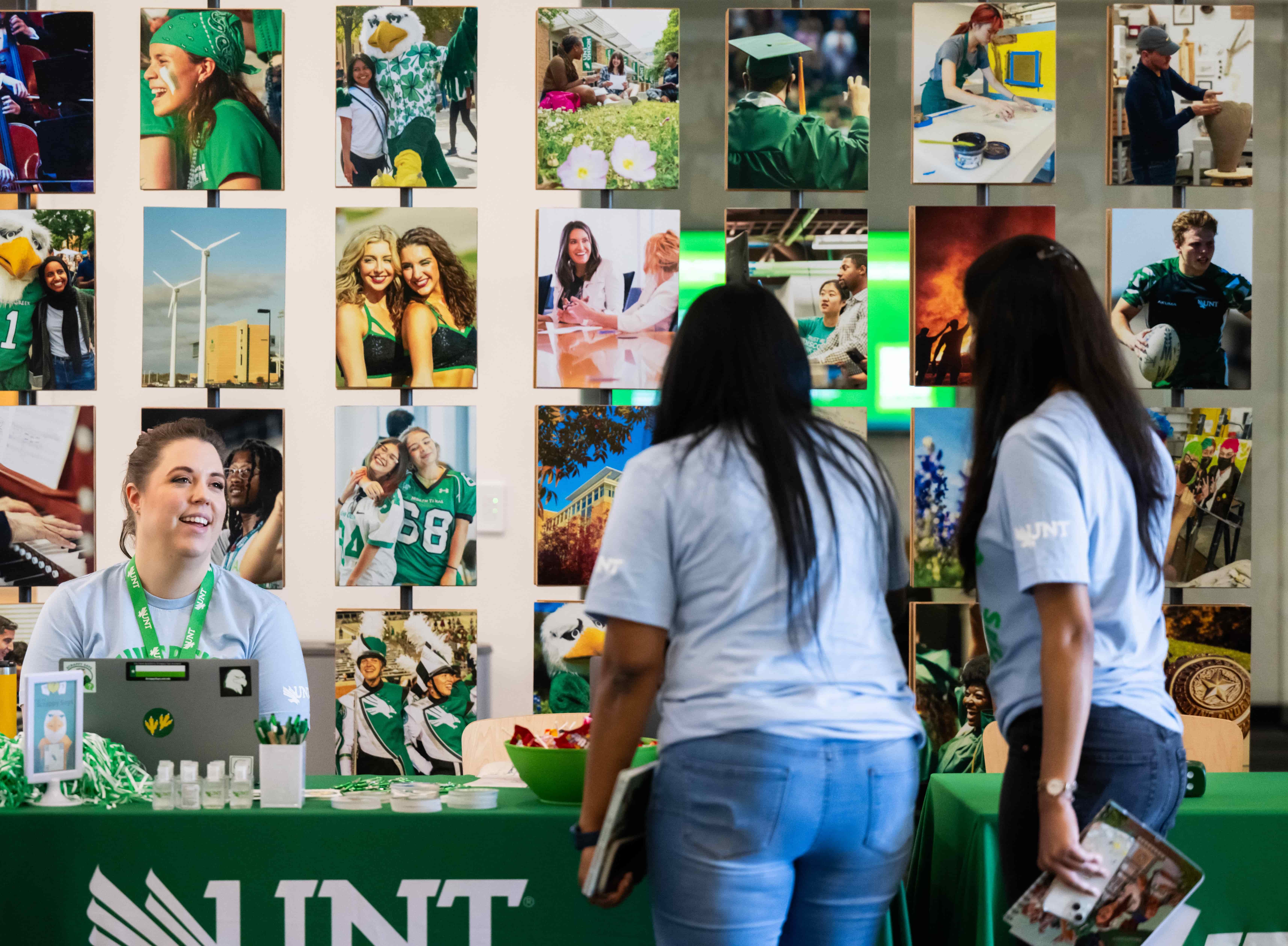 A UNT at Frisco booth during Frisco Fest. A staff member sits behind a green UNT table, smiling and talking with two students standing in front of the booth. The table displays UNT giveaways and a laptop with UNT stickers. Behind them, a wall is covered with colorful photos of campus life, students, and school spirit.