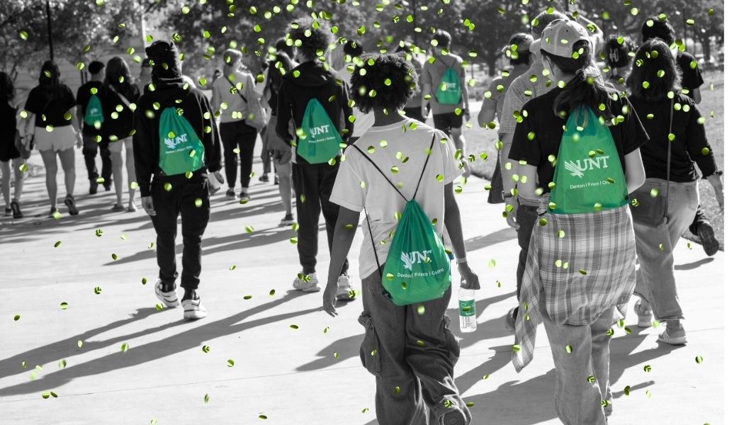 Group of students walking outdoors on campus, wearing green UNT drawstring backpacks, during an orientation event